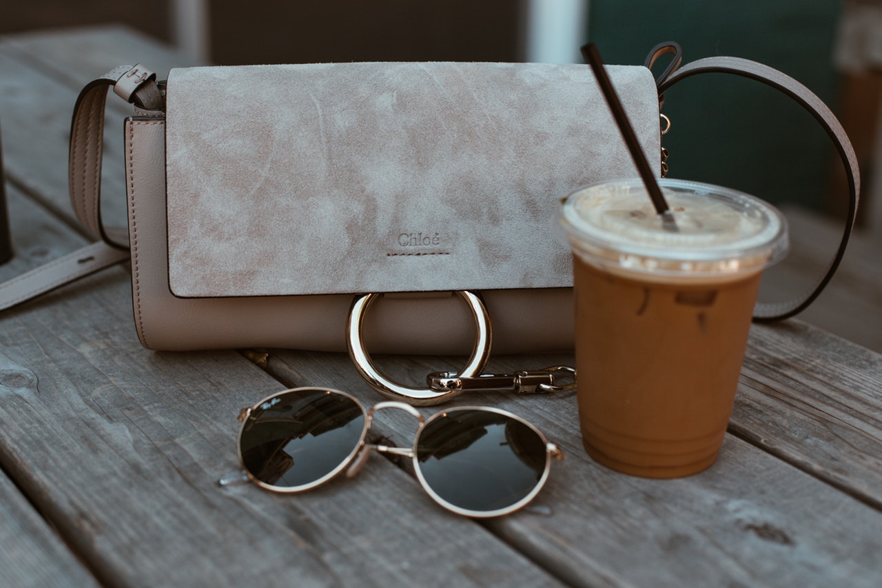 A designer handbag is sitting next to sunglasses and a coffee.