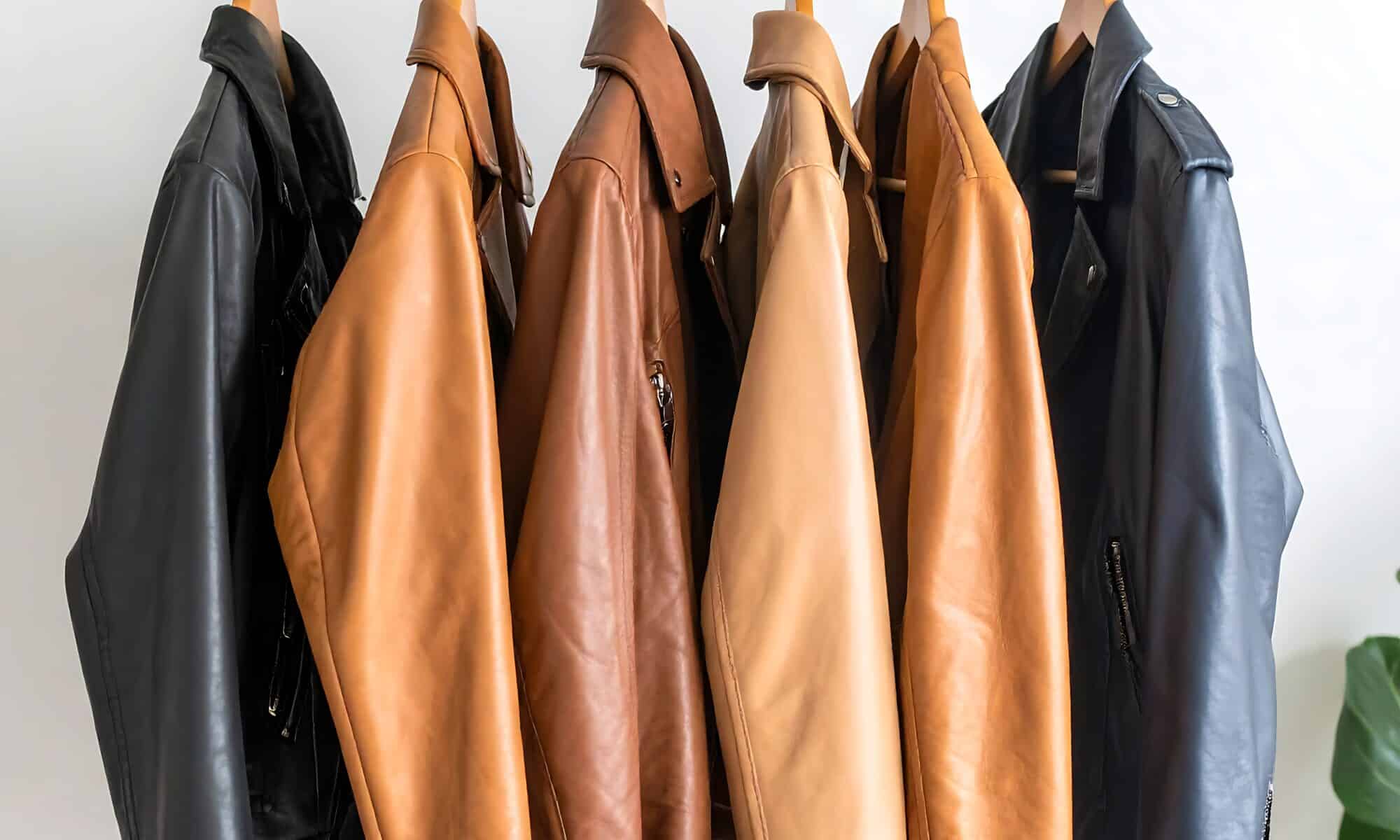 Five leather jackets in various shades of brown and black hang on a wooden rack against a white wall