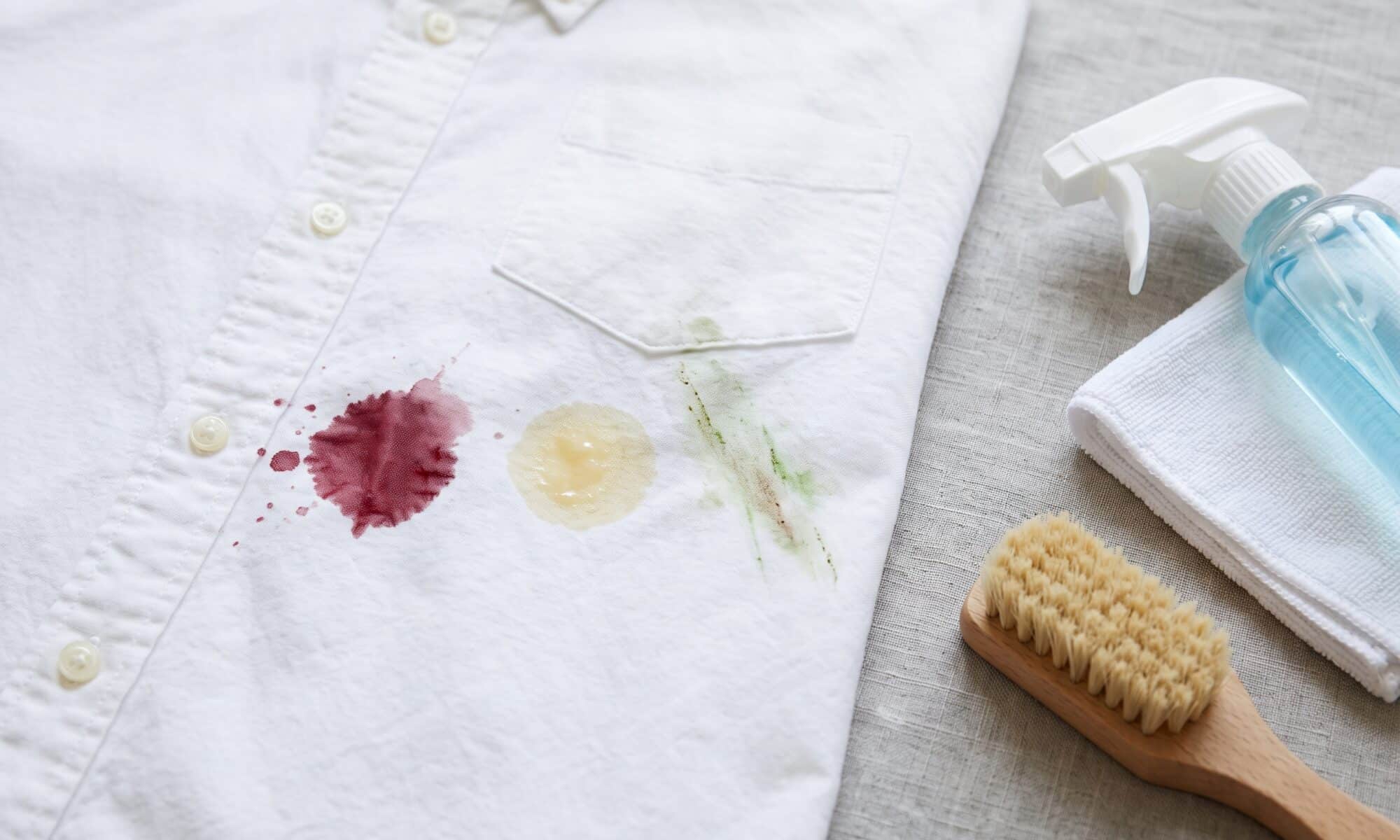 White dress shirt with wine, grease, and grass stains next to a stain remover spray, cloth, and scrub brush.